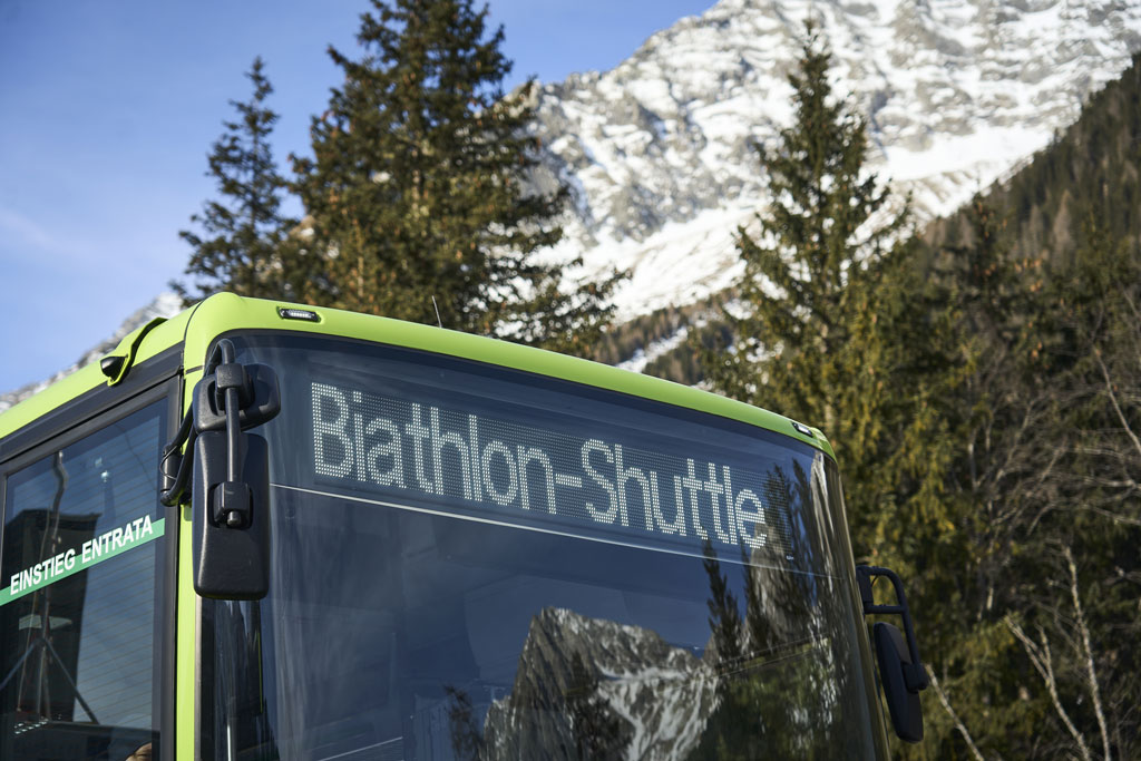 Ein grüner Linienbus mit der digitalen Anzeige „Biathlon-Shuttle“ an der Frontscheibe steht vor verschneiten Bergen und Tannenbäumen in Südtirol. An der Tür ist der Schriftzug „EINSTIEG ENTRATA“ zu sehen.