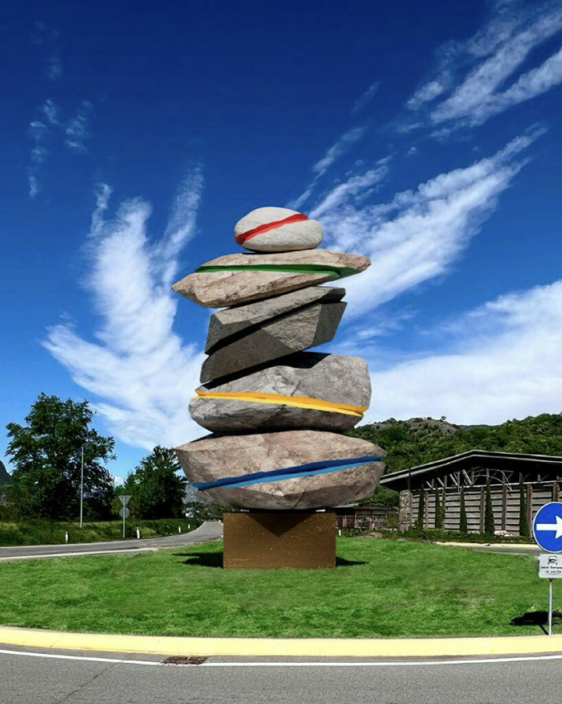 Kunstinstallation in einem Kreisverkehr: Eine vertikal balancierte Steinskulptur aus großen, unregelmäßigen Felsblöcken, die mit farbigen Linien in Rot, Grün, Schwarz, Gelb und Blau bemalt sind. Die Farben erinnern an die olympischen Ringe. Im Hintergrund: Bäume, ein modernes Gebäude und blauer Himmel mit Wolken.