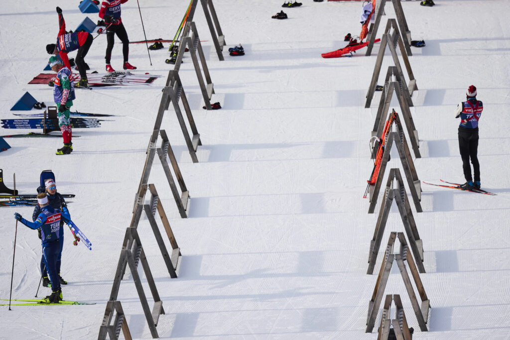 Mehrere Athlet:innen befinden sich im Skiwechselbereich auf einer verschneiten Strecke. Zwischen hölzernen Halterungen sind Skier abgelegt, einige Sportler tragen Teamwesten mit Länderaufschriften. Die Szene wirkt ruhig und vorbereitet, mit Spuren im Schnee.