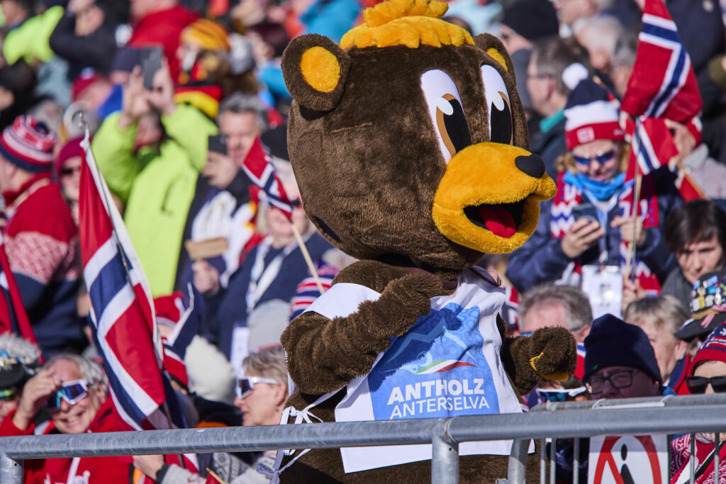 Ein großes Bären-Maskottchen mit gelber Schnauze und T-Shirt mit der Aufschrift „Antholz Anterselva“ steht vor einer jubelnden Zuschauermenge. Viele Fans in rot-blauer Kleidung schwenken norwegische Fahnen im sonnigen Biathlonstadion.